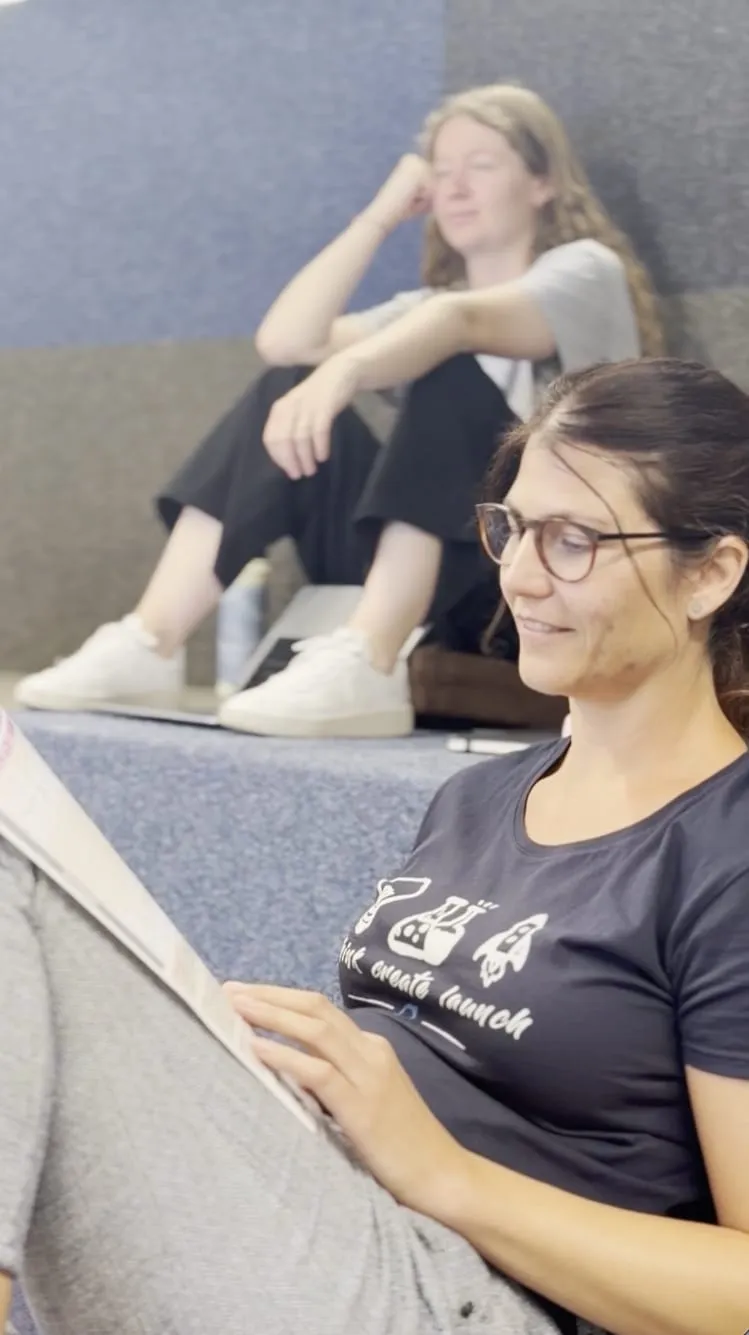 Sandra sitzt lächelnd mit den Schulungsunterlagen auf den Sitzstufen der Arena, hinter ihr Fiona