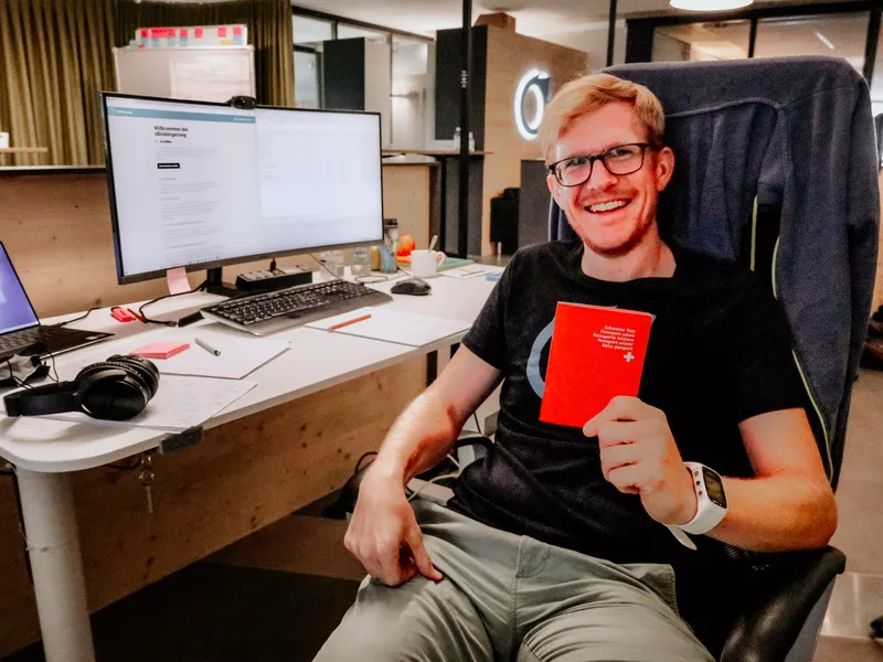 Thomas am Arbeitsplatz mit einem Schweizer Pass in der Hand, im Hintergrund das Einbürgerungsportal auf dem Bildschirm