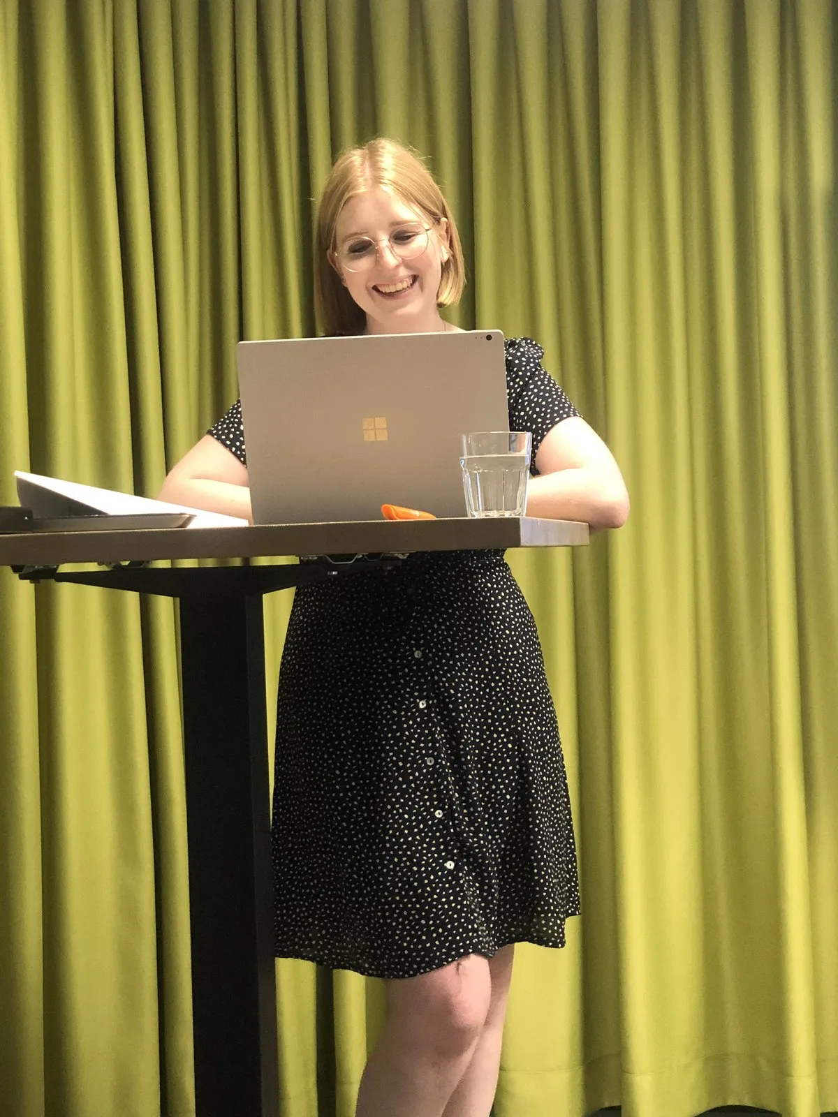 Lea präsentiert an einem Stehpult mit Laptop vor grünem Vorhang